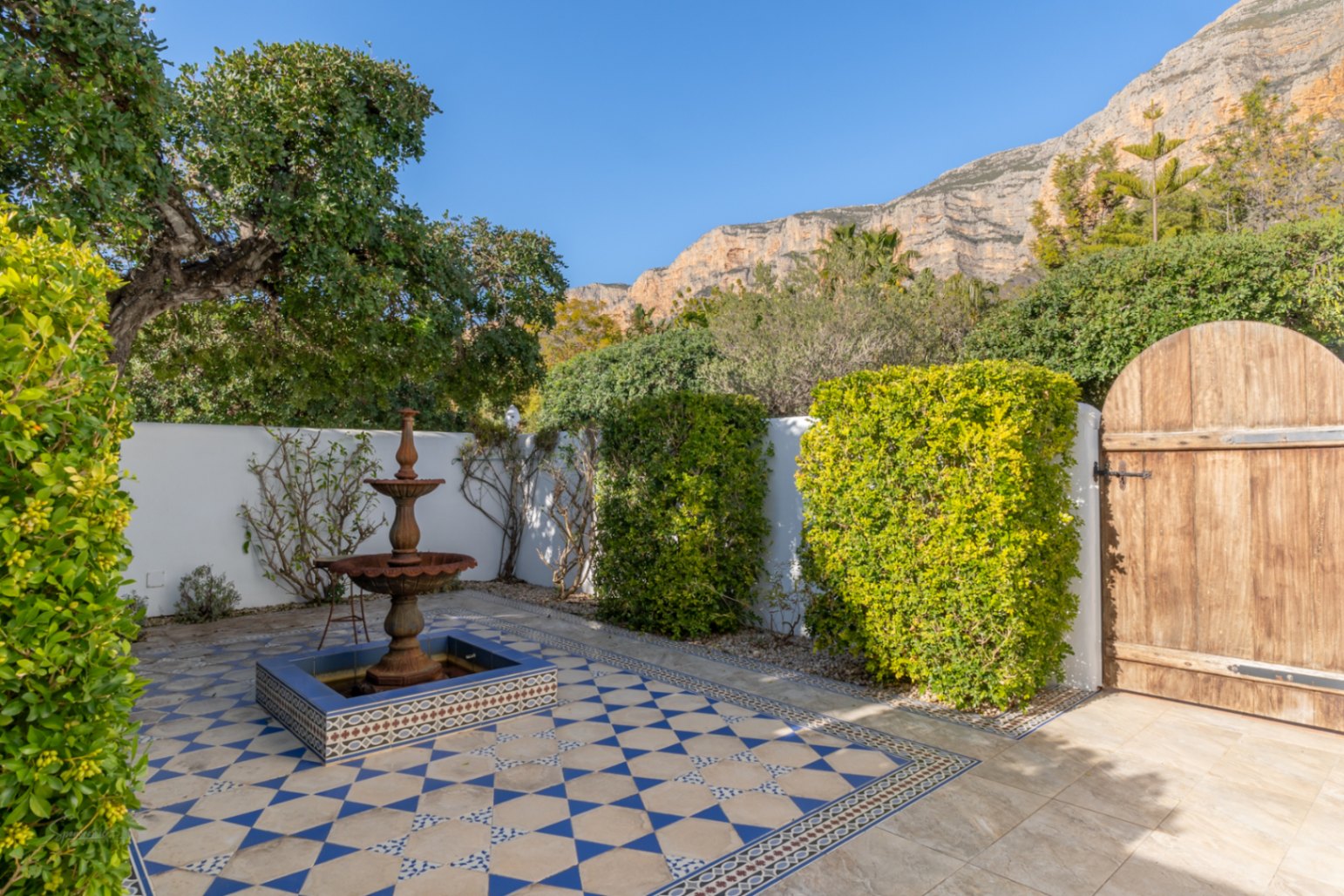 Patio de estilo andaluz con suelo de azulejos geométricos en azul y blanco, una fuente de hierro fundido y vistas a la montaña Montgó tras los muros blancos.