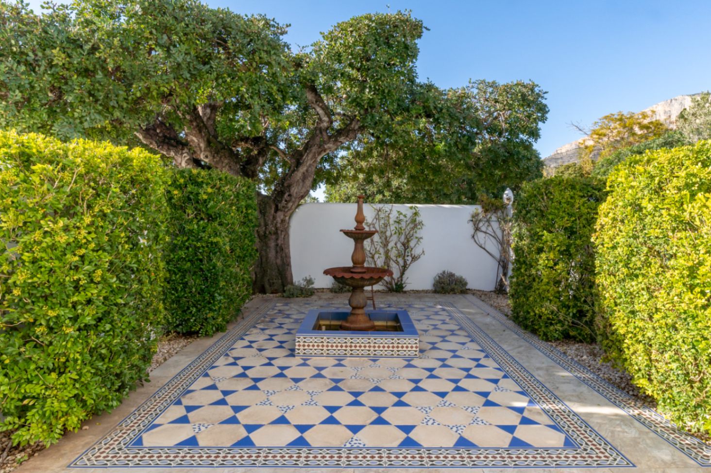 Patio de estilo mediterráneo con suelo de mosaico geométrico en azul y blanco, presidido por una fuente de hierro fundido y rodeado de setos frondosos.