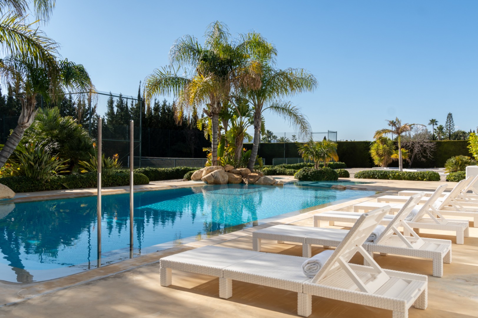 Piscina de 70m² con zona de hamacas blancas, rodeada de palmeras y vegetación mediterránea, con una pista de tenis privada visible al fondo bajo un cielo despejado.