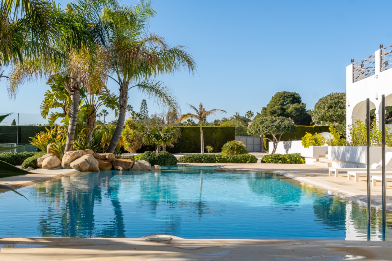 Piscina de diseño con entrada gradual, rodeada de jardines tropicales, palmeras y rocas decorativas, junto a la naya de arcos blancos y la pista de tenis.