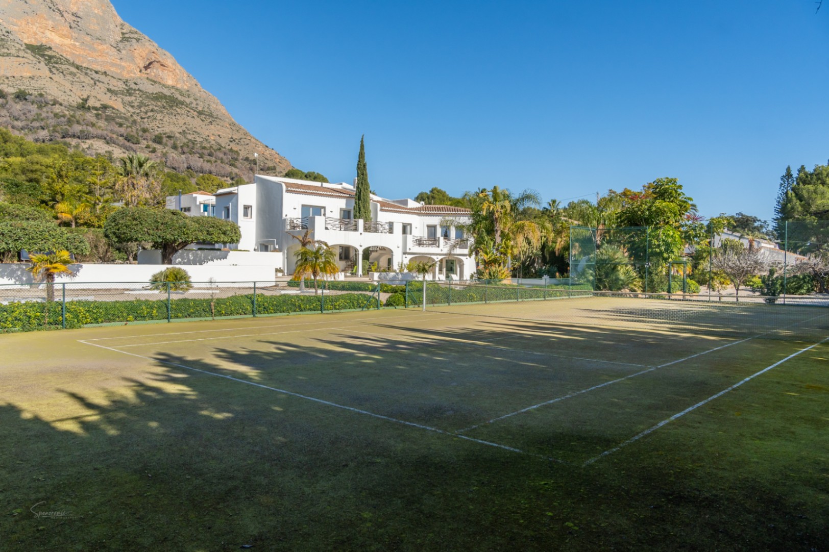 Pista de tenis privada con superficie verde y vallado perimetral, situada frente a la villa de estilo ibicenco con el macizo del Montgó al fondo.