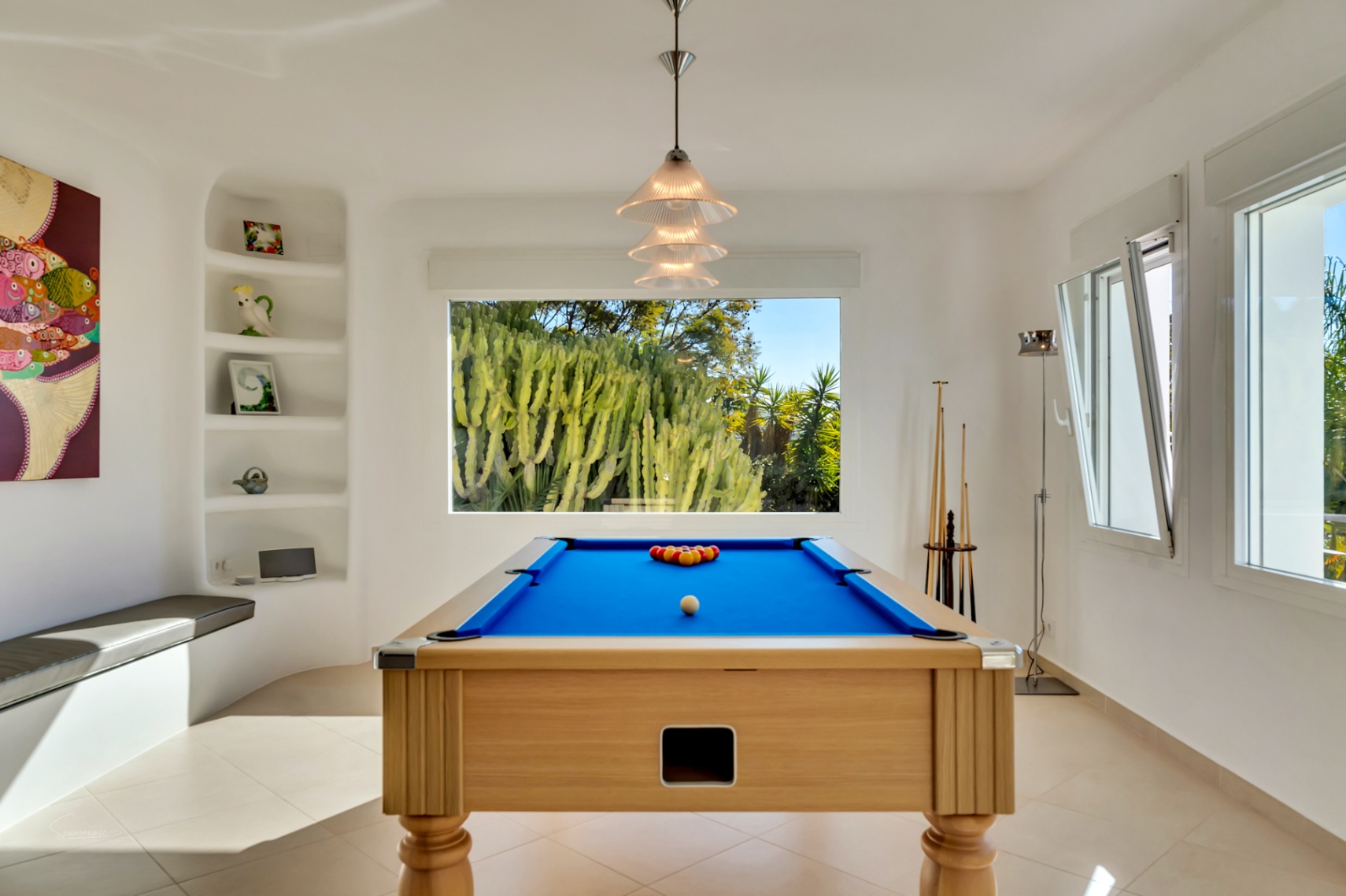 Sala de juegos con mesa de billar de madera y tapete azul, iluminada por luz natural y vistas a un jardín de cactus a través de un gran ventanal.