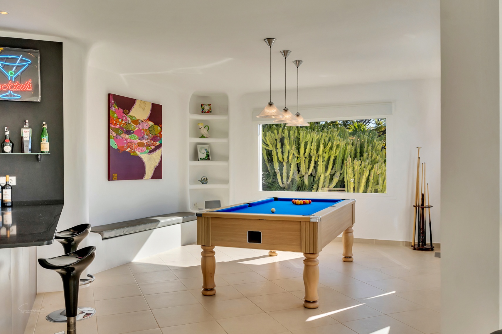 Sala de juegos con mesa de billar, zona de bar integrada con taburetes y ventanal con vistas a cactus. El diseño presenta paredes blancas curvas de estilo ibicenco y suelos cerámicos.