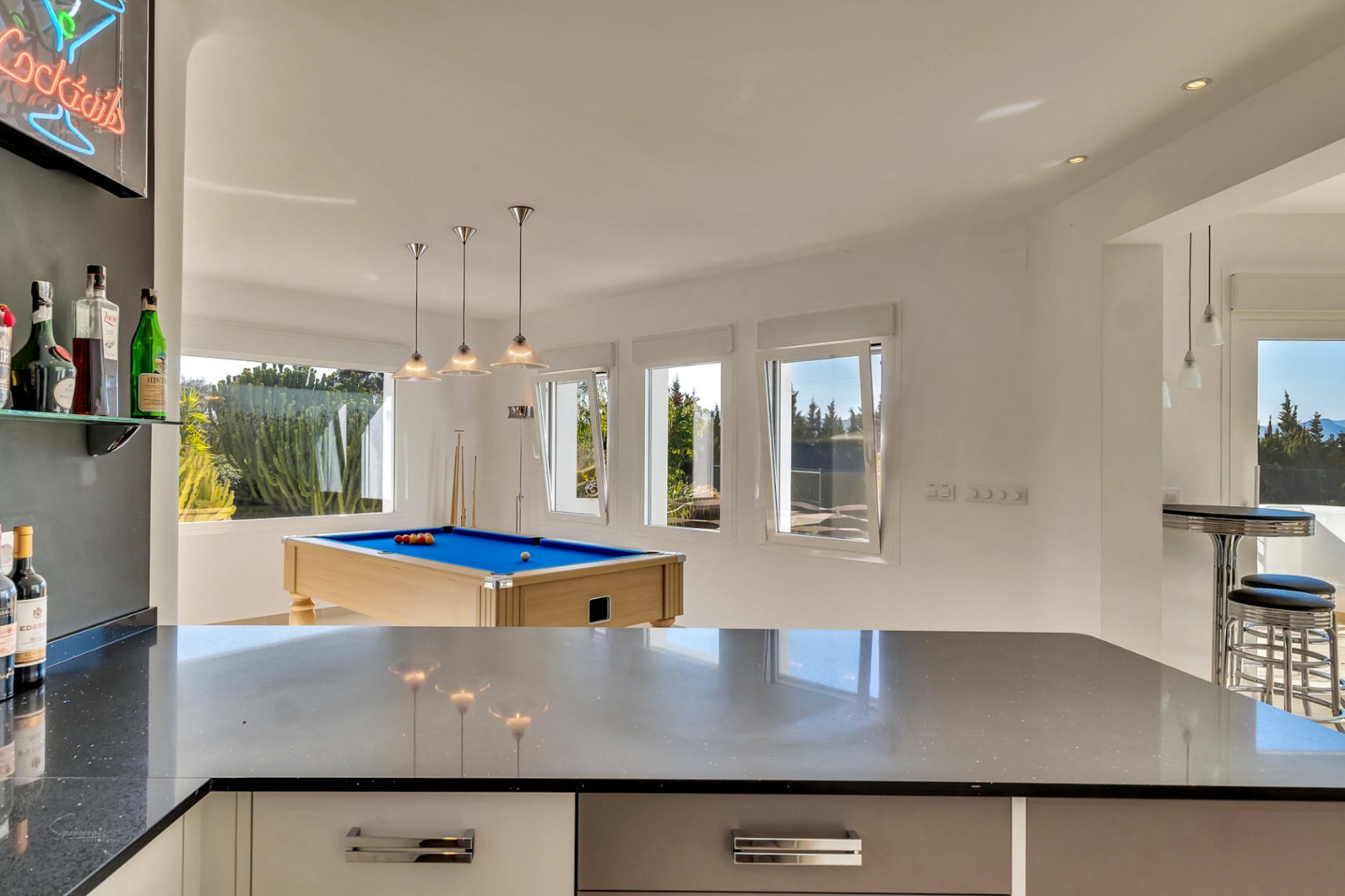 Sala de ocio con mesa de billar de tapete azul, barra de bar con encimera oscura y amplios ventanales que aportan abundante luz natural.