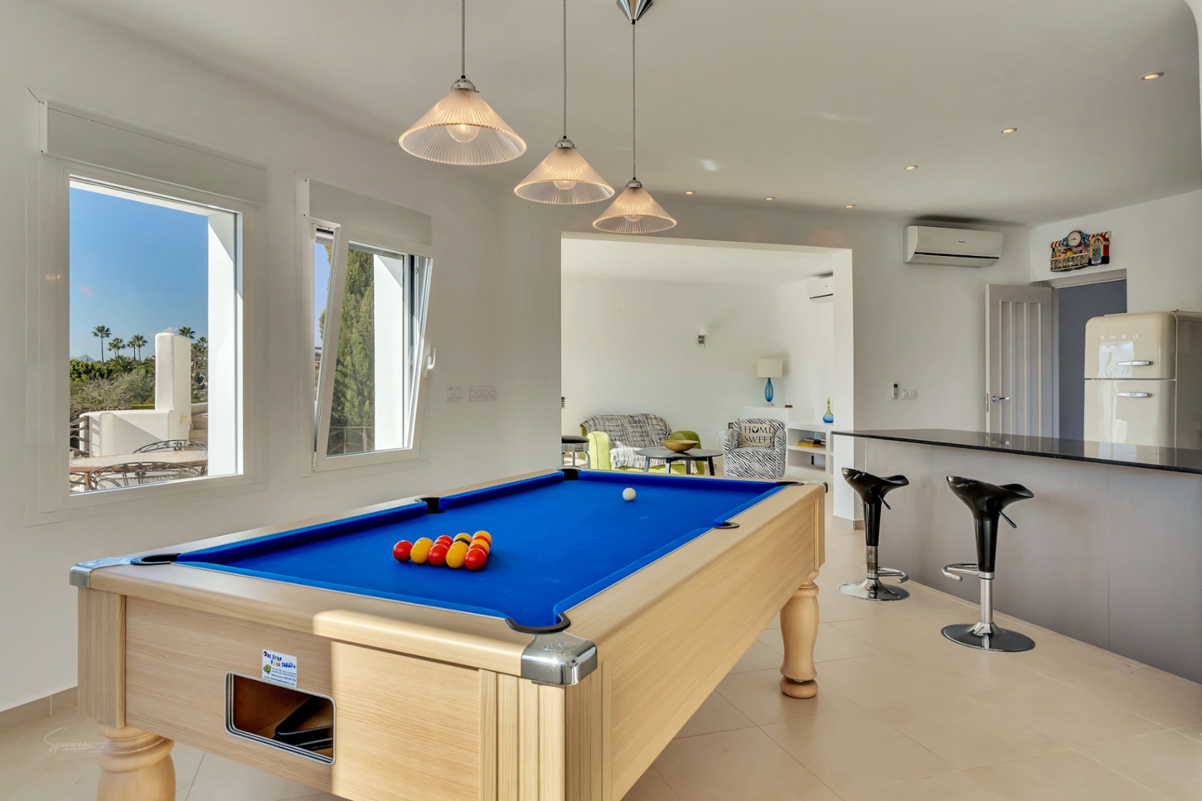 Sala de ocio con mesa de billar profesional, barra de cocina con taburetes y frigorífico de estilo retro. Los ventanales aportan luz natural y vistas al entorno mediterráneo.