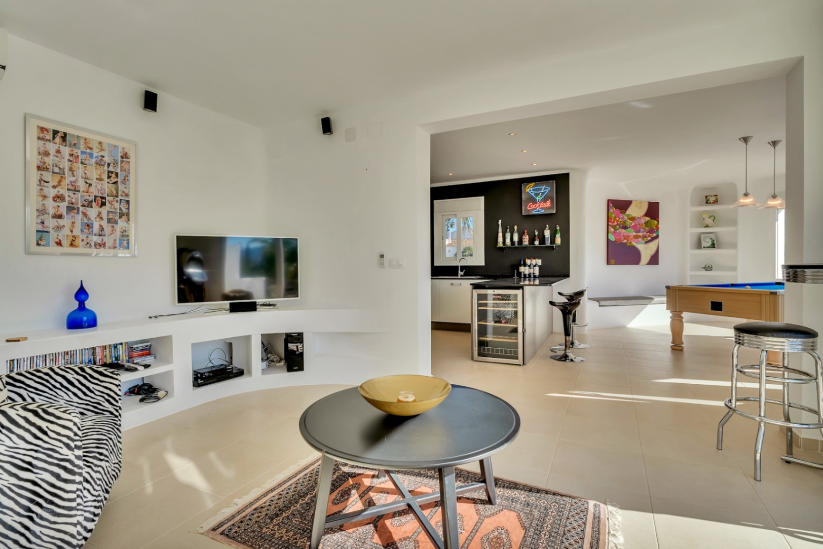 Salón de concepto abierto con mueble de TV curvo integrado, zona de bar con vinoteca y un área de entretenimiento con mesa de billar y suelos cerámicos.