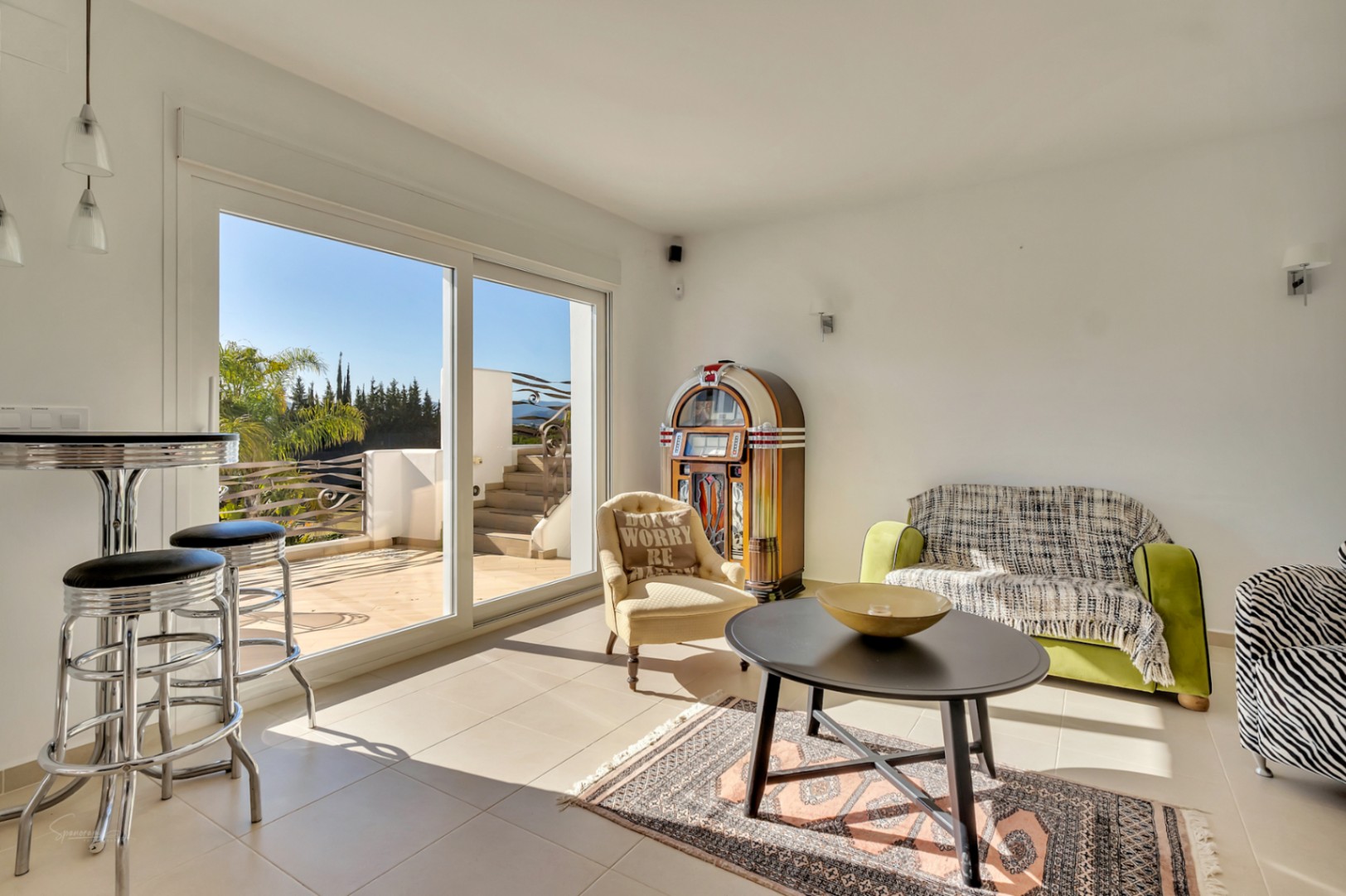 Salón luminoso con una gramola clásica, zona de estar con sofás y mesa alta con taburetes cromados, conectado a una terraza exterior mediante ventanales.