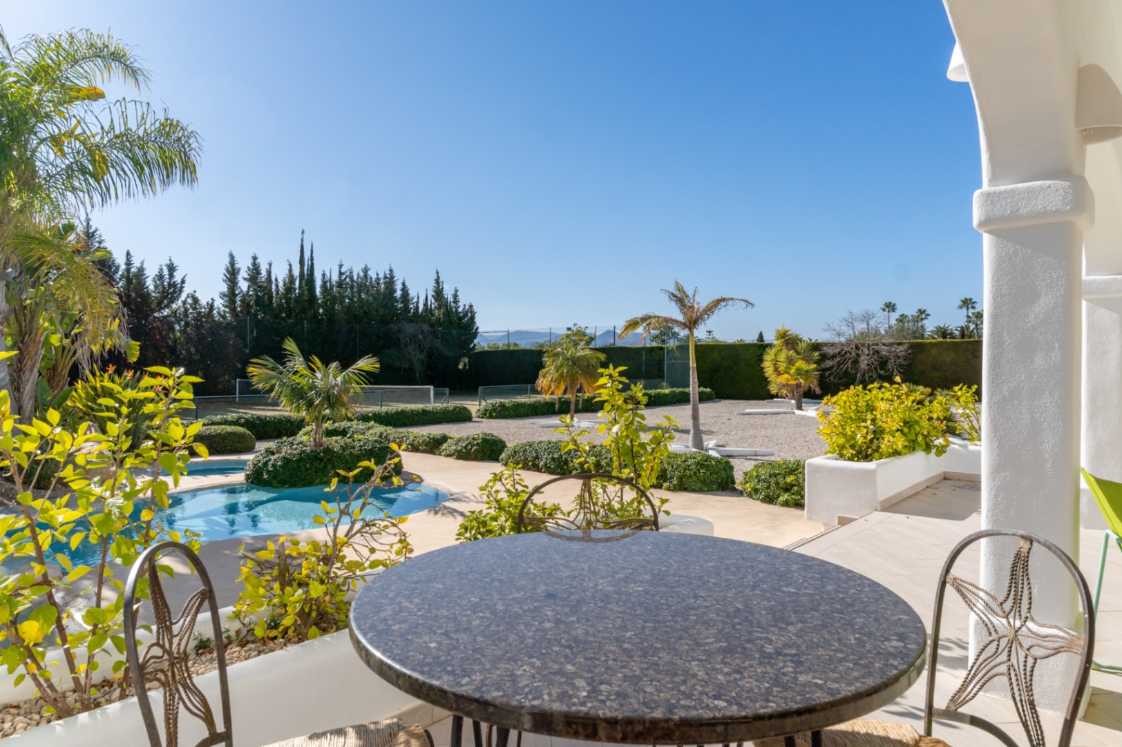 Terraza cubierta con mesa de piedra y vistas directas a la piscina, el jardín mediterráneo y la pista de tenis privada bajo un cielo despejado.