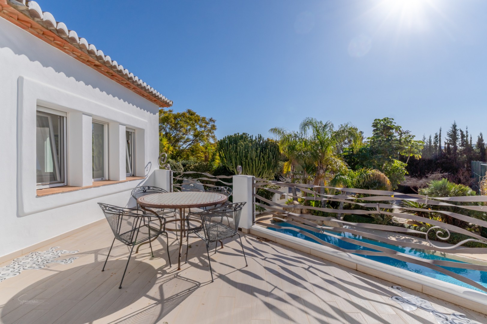 Terraza superior con barandilla de hierro artesanal, mesa de mosaico y vistas despejadas a la piscina y al jardín mediterráneo bajo el sol.