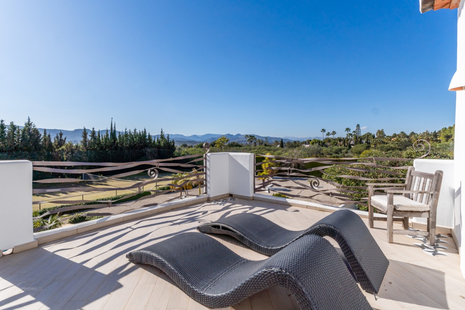 Terraza superior con barandillas de hierro onduladas, tumbonas ergonómicas y vistas despejadas hacia la pista de tenis y el valle montañoso.