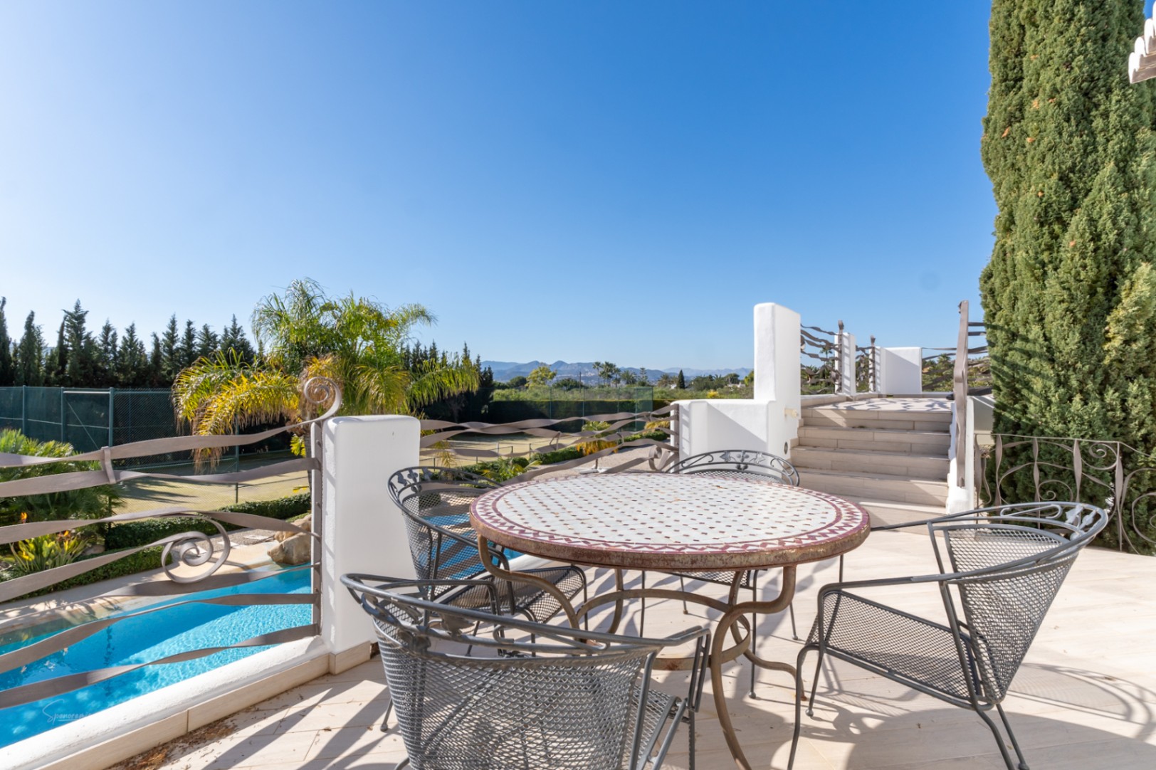 Terraza superior con mesa de mosaico y barandillas de hierro, con vistas directas a la piscina, la pista de tenis y el valle del Montgó.