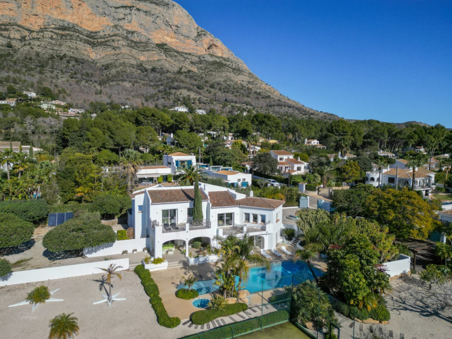 Vista aérea de la villa estilo Ibiza con fachada blanca y tejados de terracota, destacando la piscina privada, pista de tenis y el telón de fondo del Montgó.