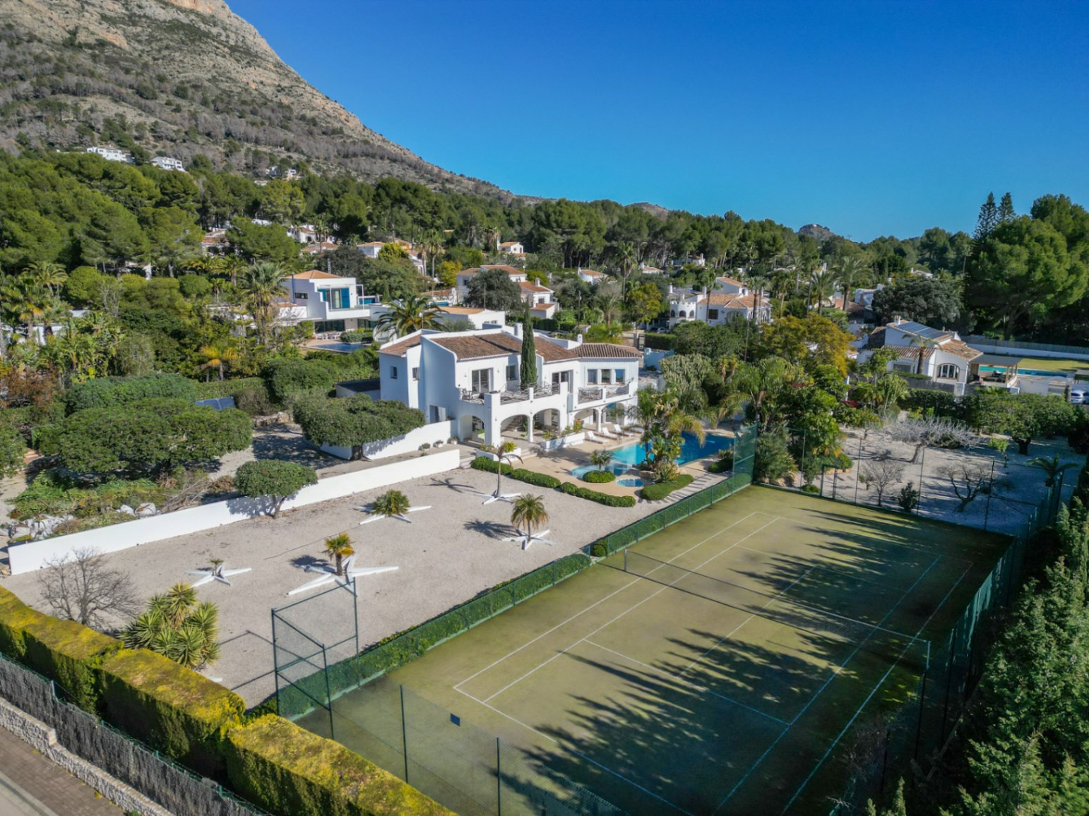 Vista aérea de la villa estilo Ibiza con pista de tenis privada, piscina con jacuzzi y jardín mediterráneo bajo la falda de la montaña Montgó.