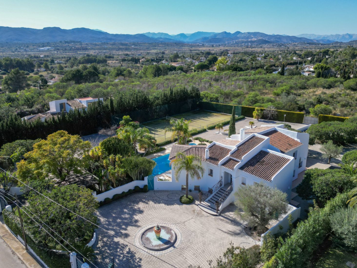 Vista aérea de la villa estilo Ibiza con su entrada circular pavimentada, fuente central, piscina privada, pista de tenis y vistas panorámicas al valle y al Montgó.