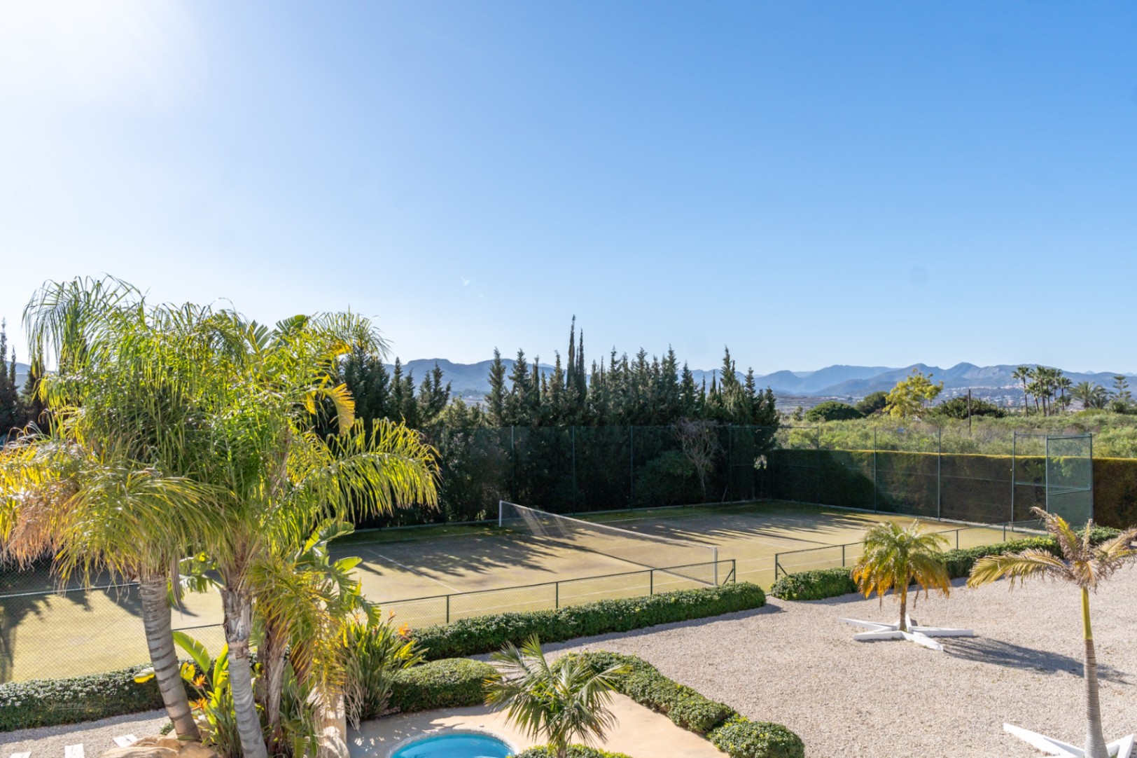 Vista elevada de la cancha de tenis privada rodeada de setos maduros y palmeras, con el valle y las montañas al fondo bajo un cielo despejado.