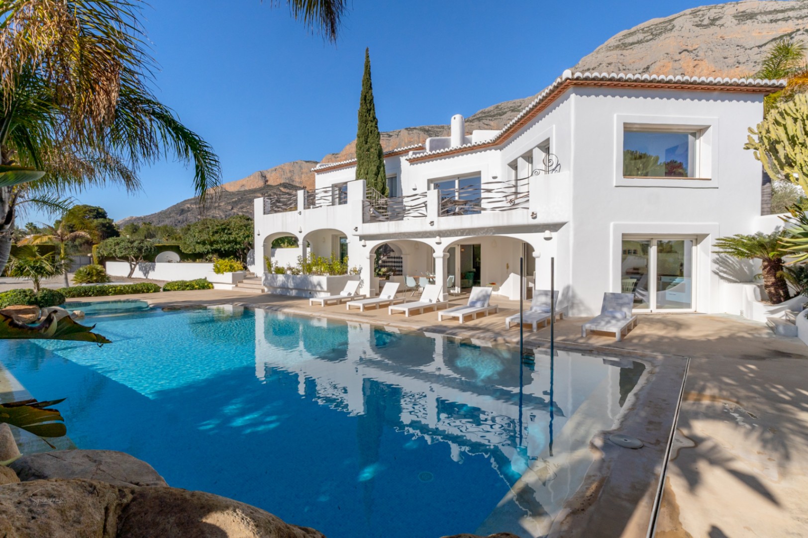 Vista exterior de la villa blanca estilo Ibiza con naya de arcos, amplia piscina de aguas cristalinas y el macizo del Montgó al fondo.