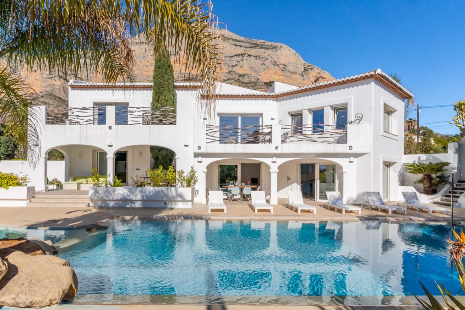 Vista exterior de la villa estilo Ibiza con piscina de 70m², amplias nayas arqueadas y terrazas con barandillas de hierro, bajo la imponente montaña Montgó.