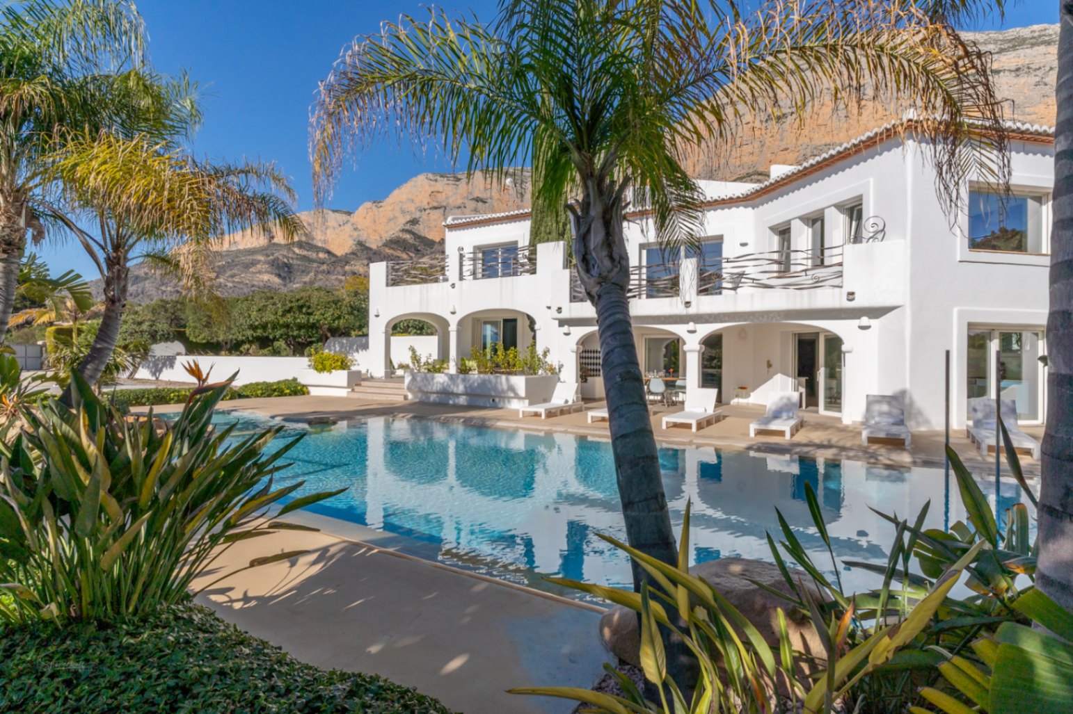 Vista exterior de la villa estilo Ibiza con piscina de 70m², rodeada de palmeras y vegetación tropical, bajo el imponente macizo del Montgó.