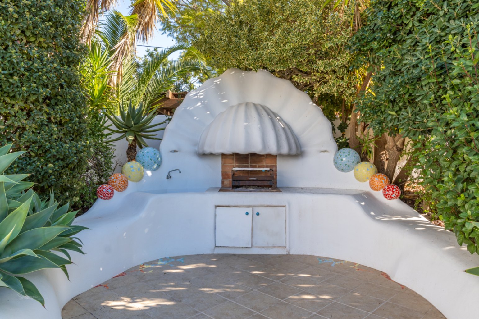 Zona de barbacoa de obra con diseño escultural en forma de concha, rodeada de vegetación exuberante y decorada con esferas de mosaico sobre un suelo de gres cerámico.