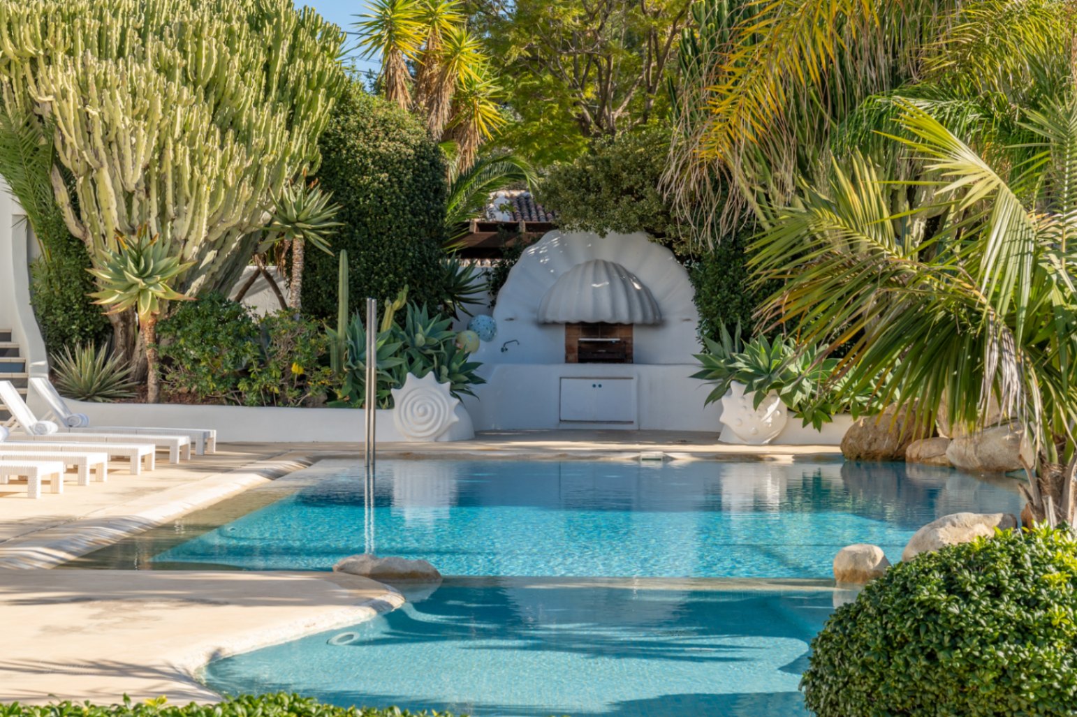 Zona de piscina con barbacoa blanca de diseño orgánico, rodeada de vegetación mediterránea, cactus de gran tamaño y tumbonas blancas sobre suelo de piedra clara.