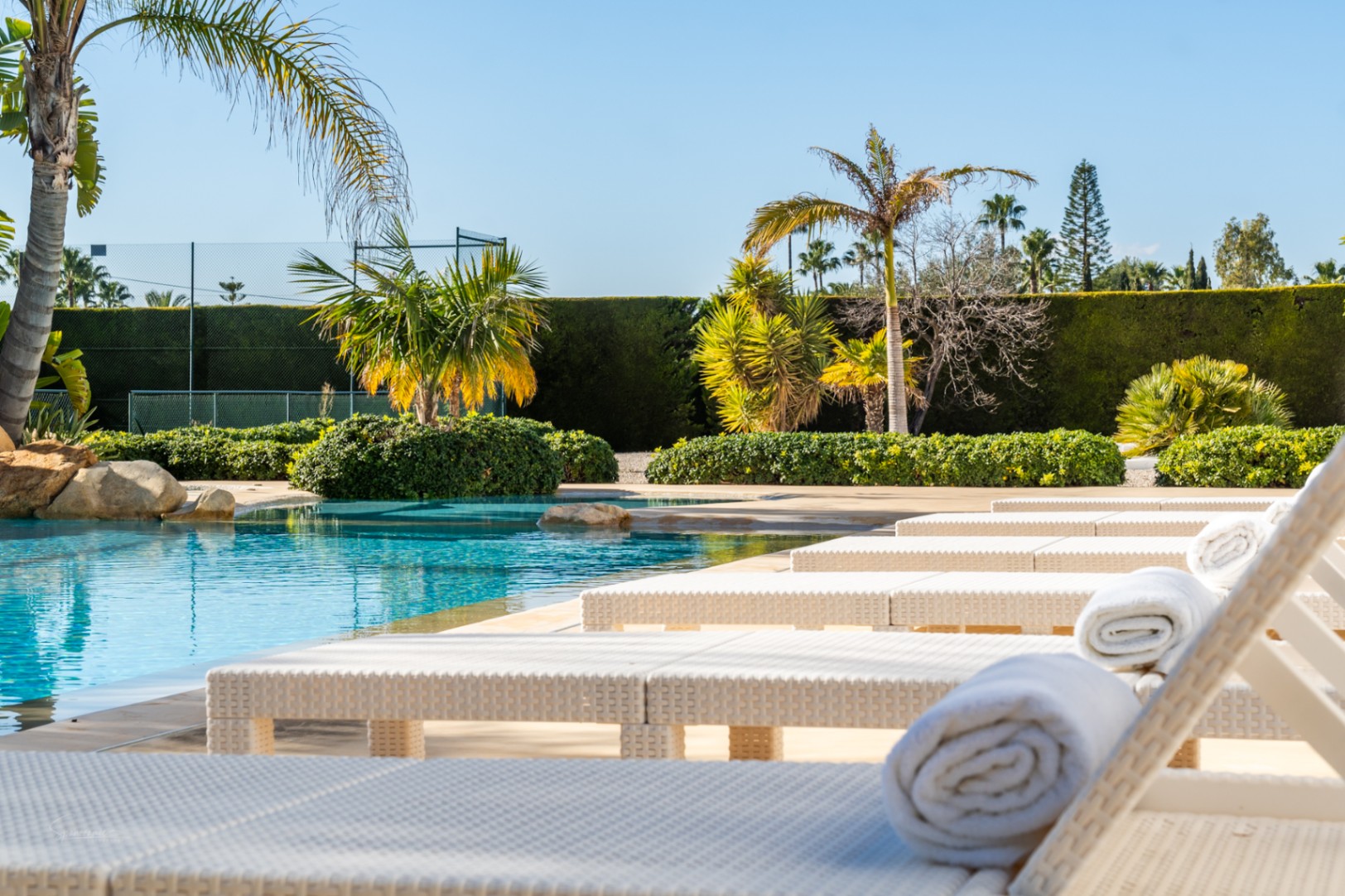 Zona de piscina con tumbonas de ratán blanco y toallas, junto a un jardín mediterráneo con palmeras y una pista de tenis privada al fondo.