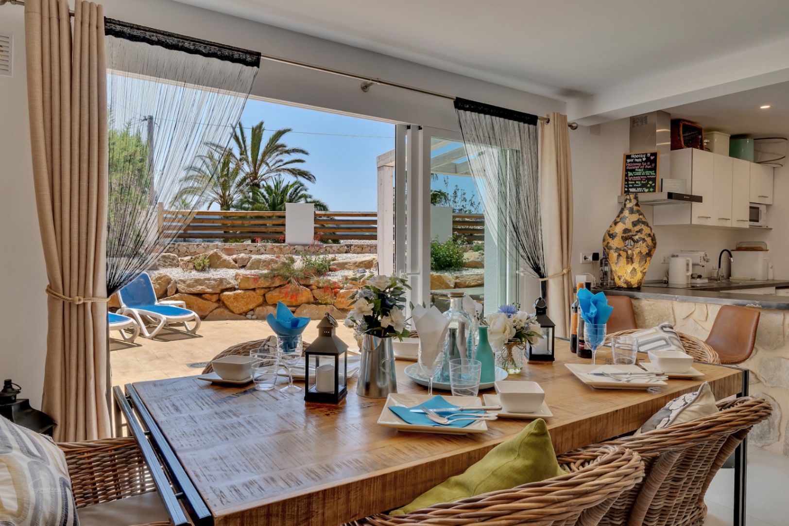 Comedor luminoso con mesa rústica y acceso a terraza con tumbonas y jardín mediterráneo.