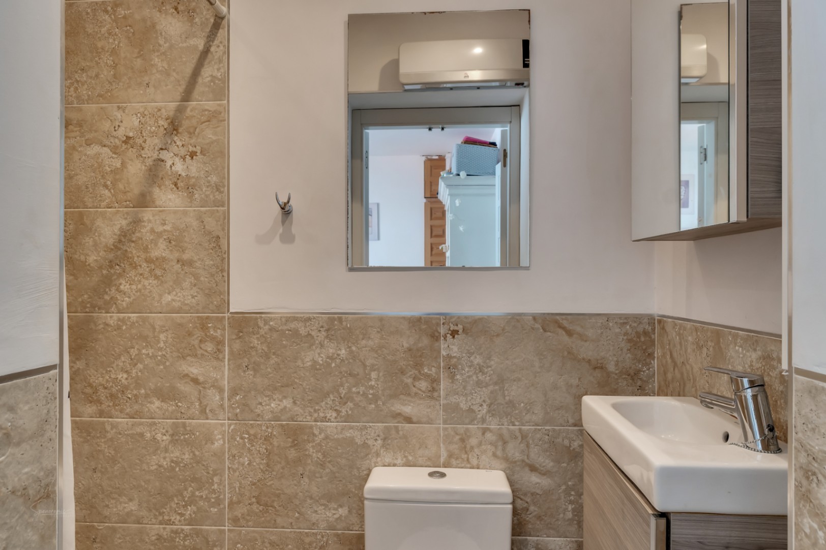 Detalle de baño con azulejos color crema y espejo que refleja otra estancia.