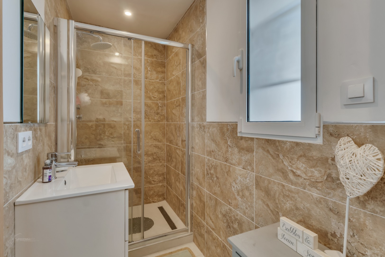 Detalle del baño con ducha de mampara corredera y lavabo moderno, revestido con azulejos de color tierra.