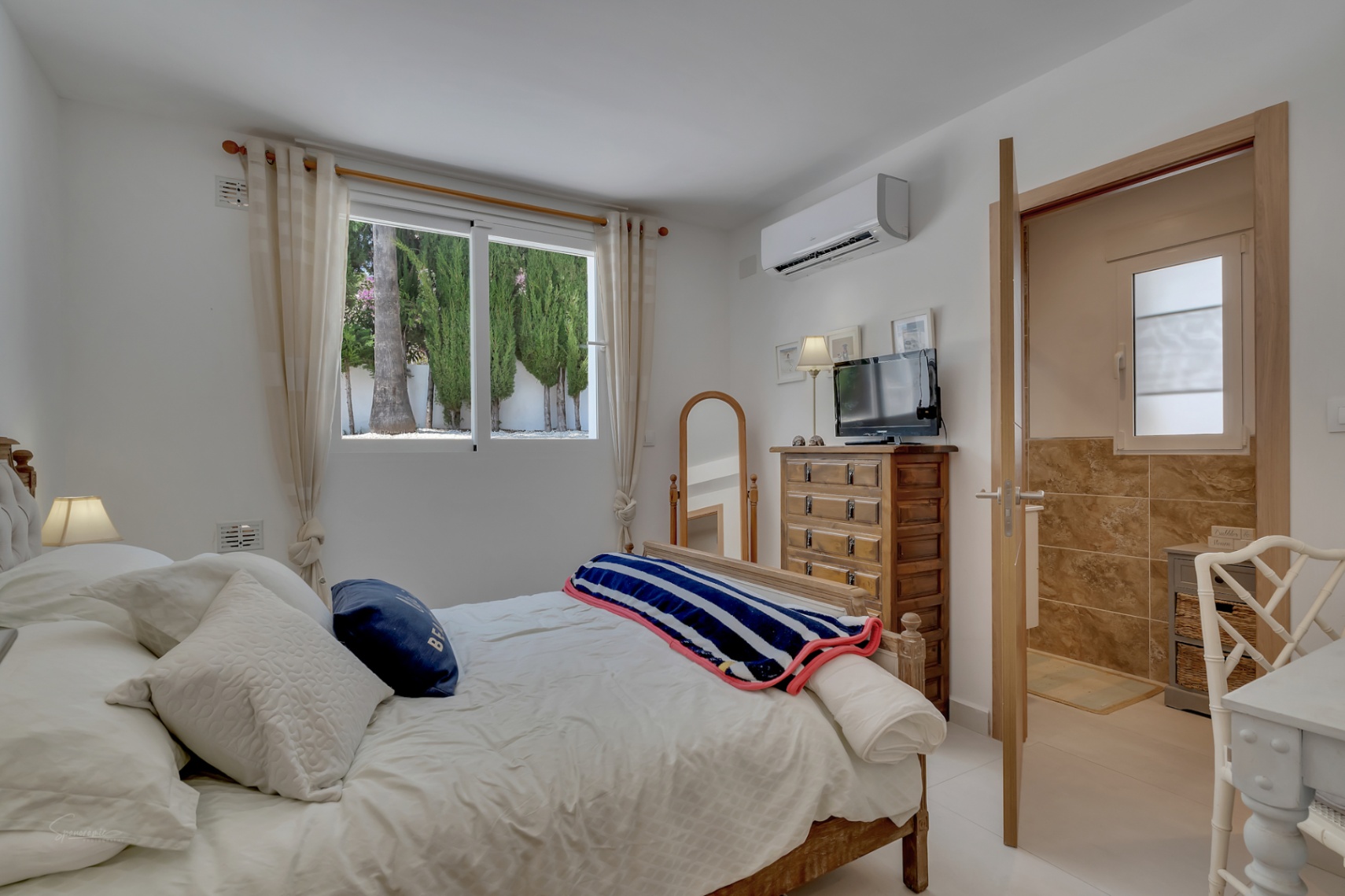 Dormitorio luminoso con cama de madera, cómoda rústica y baño en suite con azulejos color tierra.