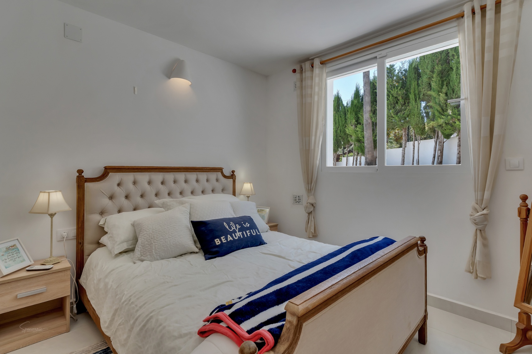 Dormitorio luminoso con cama de madera y cabecero tapizado. Vista a los cipreses desde la ventana.