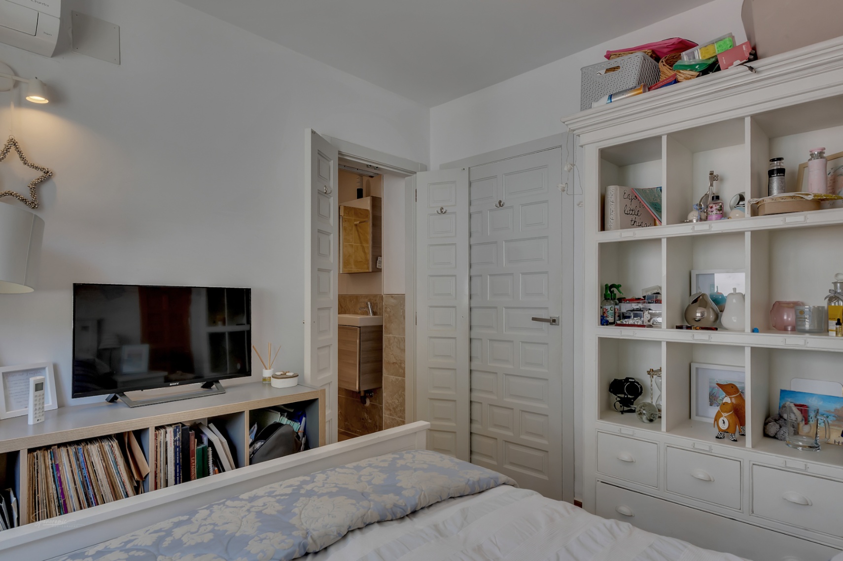 Dormitorio luminoso con estantería blanca y acceso al baño en suite con detalles en madera.