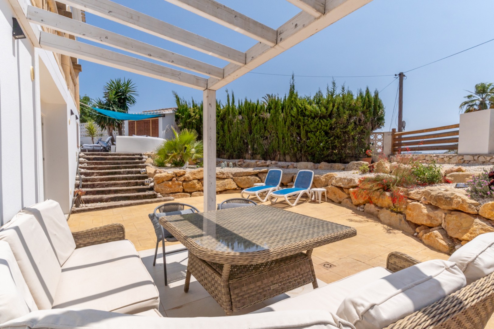 Terraza con pérgola blanca, mobiliario de ratán y vistas al jardín con tumbonas azules.