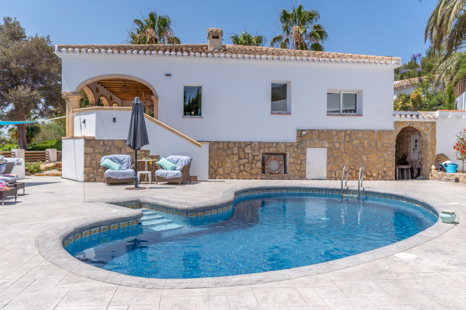 Vista de la piscina con mosaicos azules y fachada de piedra rústica de la villa en Jávea.