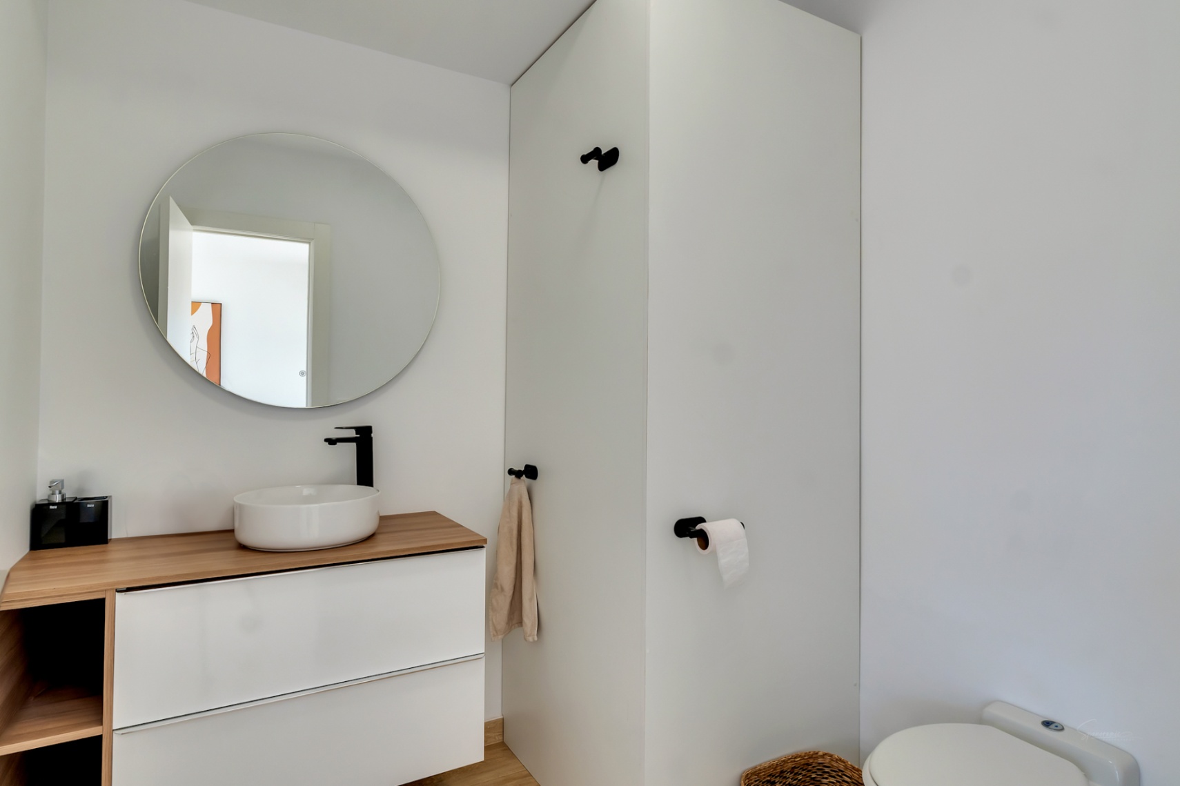 Baño moderno con lavabo sobre encimera de madera, grifería negra mate y espejo circular. El diseño minimalista incluye paredes blancas y accesorios contemporáneos.