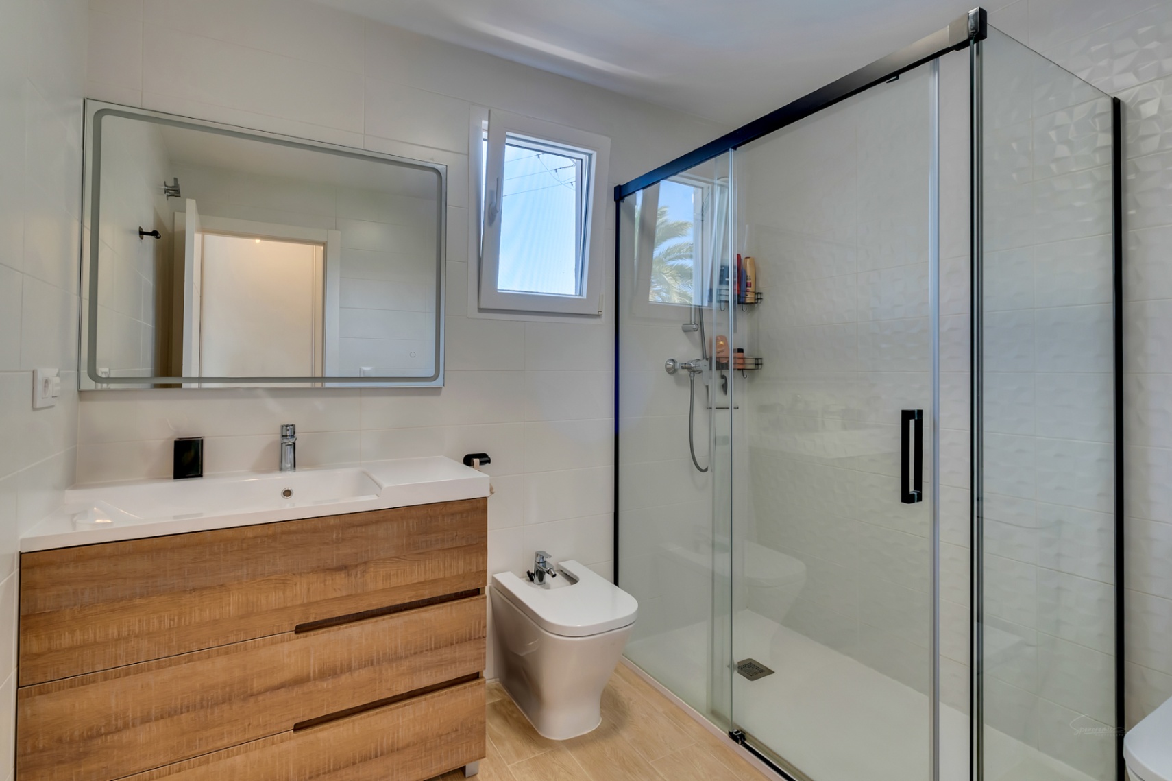 Baño moderno con mueble de lavabo en madera, bidet y amplia ducha de obra con mampara de perfilería negra y azulejos blancos texturizados.