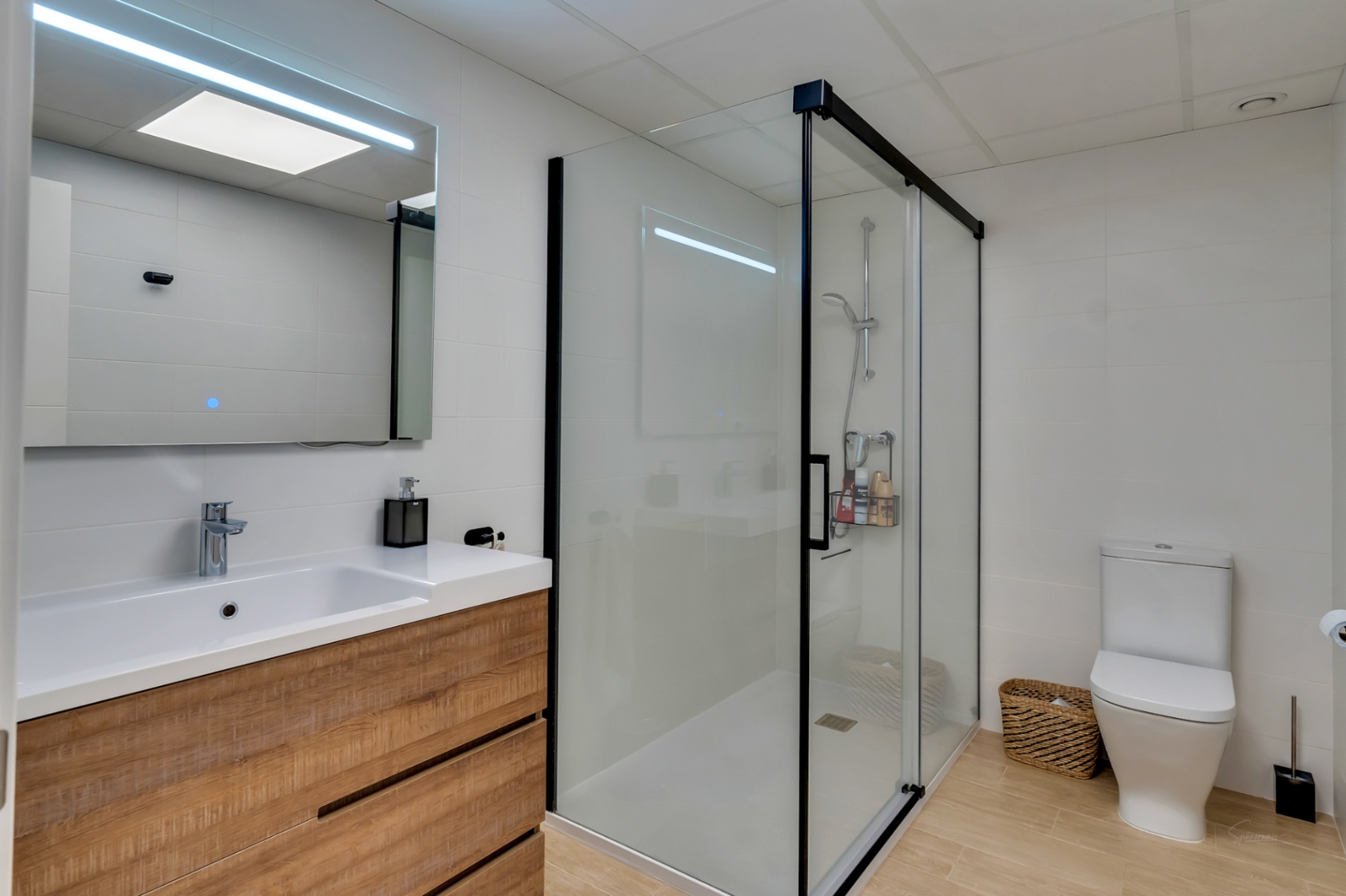 Baño moderno con mueble de madera, lavabo integrado y espejo con iluminación LED. Incluye una amplia ducha con perfilería negra y suelos de gres efecto madera.