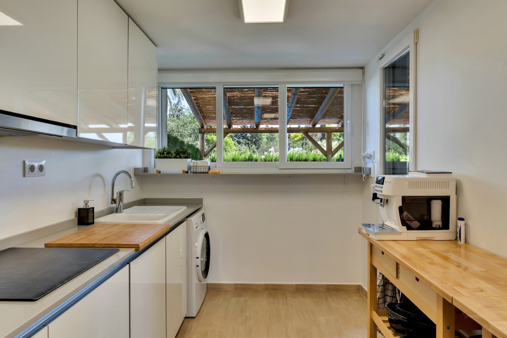 Cocina moderna con armarios blancos brillantes, zona de lavandería integrada y un gran ventanal con vistas a la pérgola de madera del patio privado.
