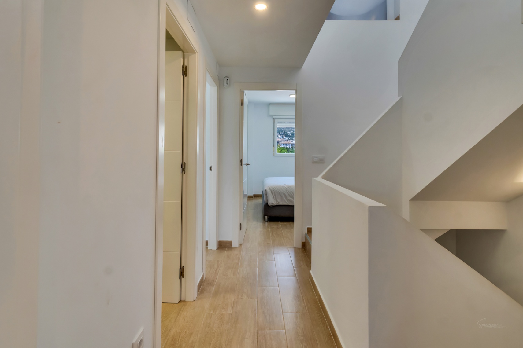 Distribuidor con suelos cerámicos de imitación madera y escalera blanca minimalista. Al fondo se aprecia un dormitorio luminoso con vistas despejadas al entorno.