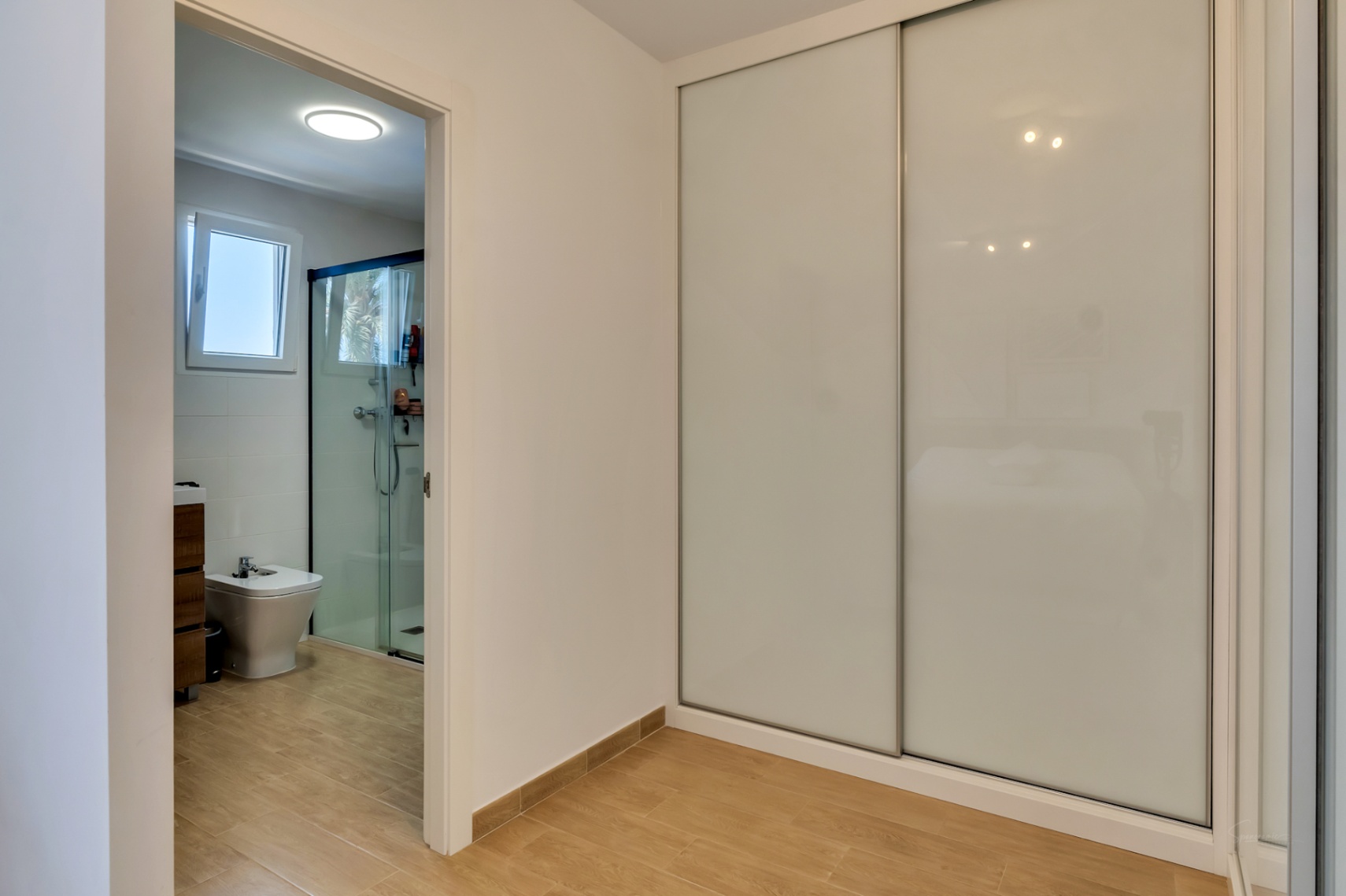 Dormitorio con amplios armarios empotrados de puertas correderas blancas y acceso directo a un baño en suite con ducha de obra y acabados modernos.