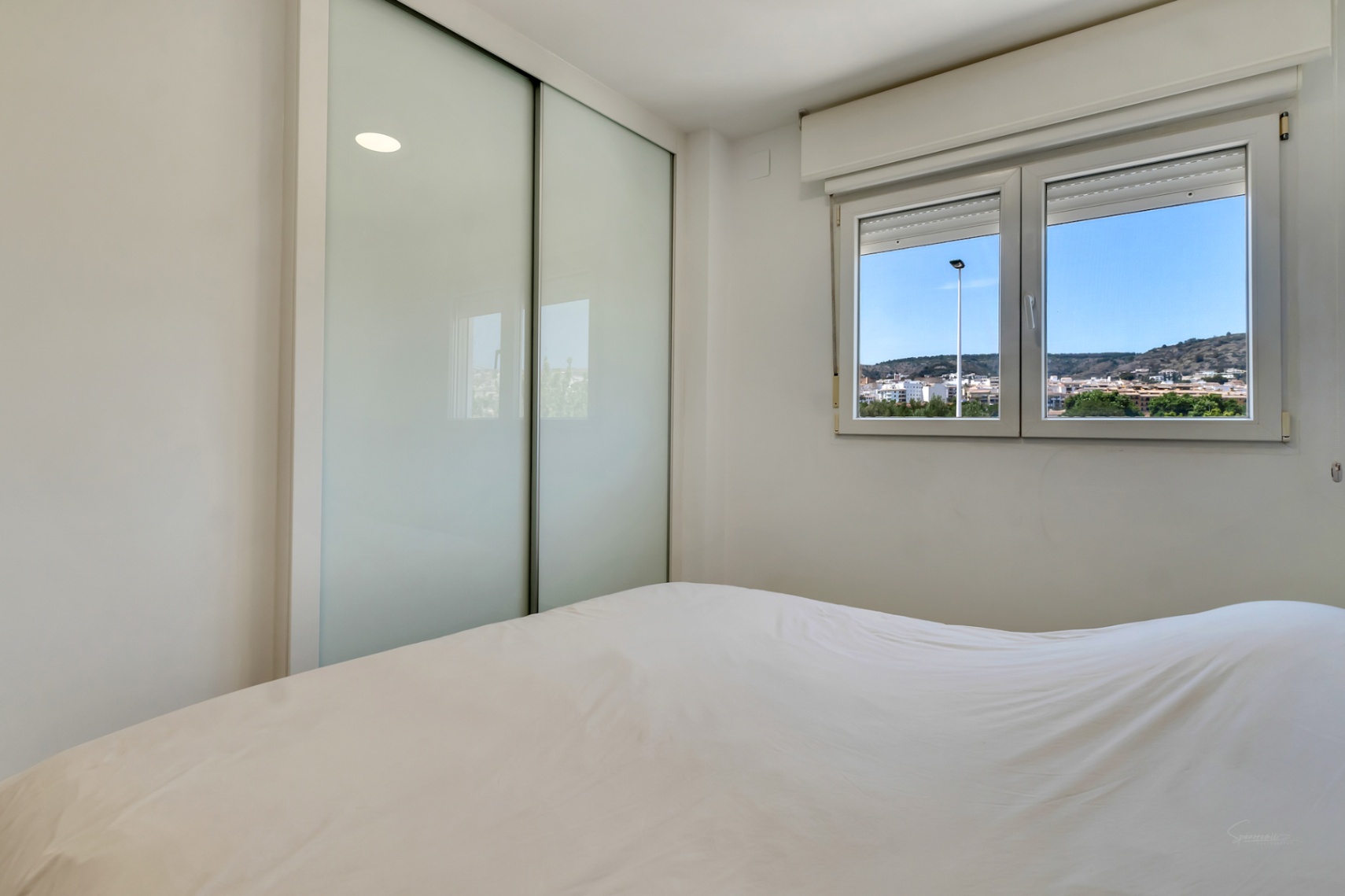Dormitorio luminoso con armario empotrado de puertas correderas en cristal translúcido y ventana de PVC con vistas despejadas al entorno de Jávea.