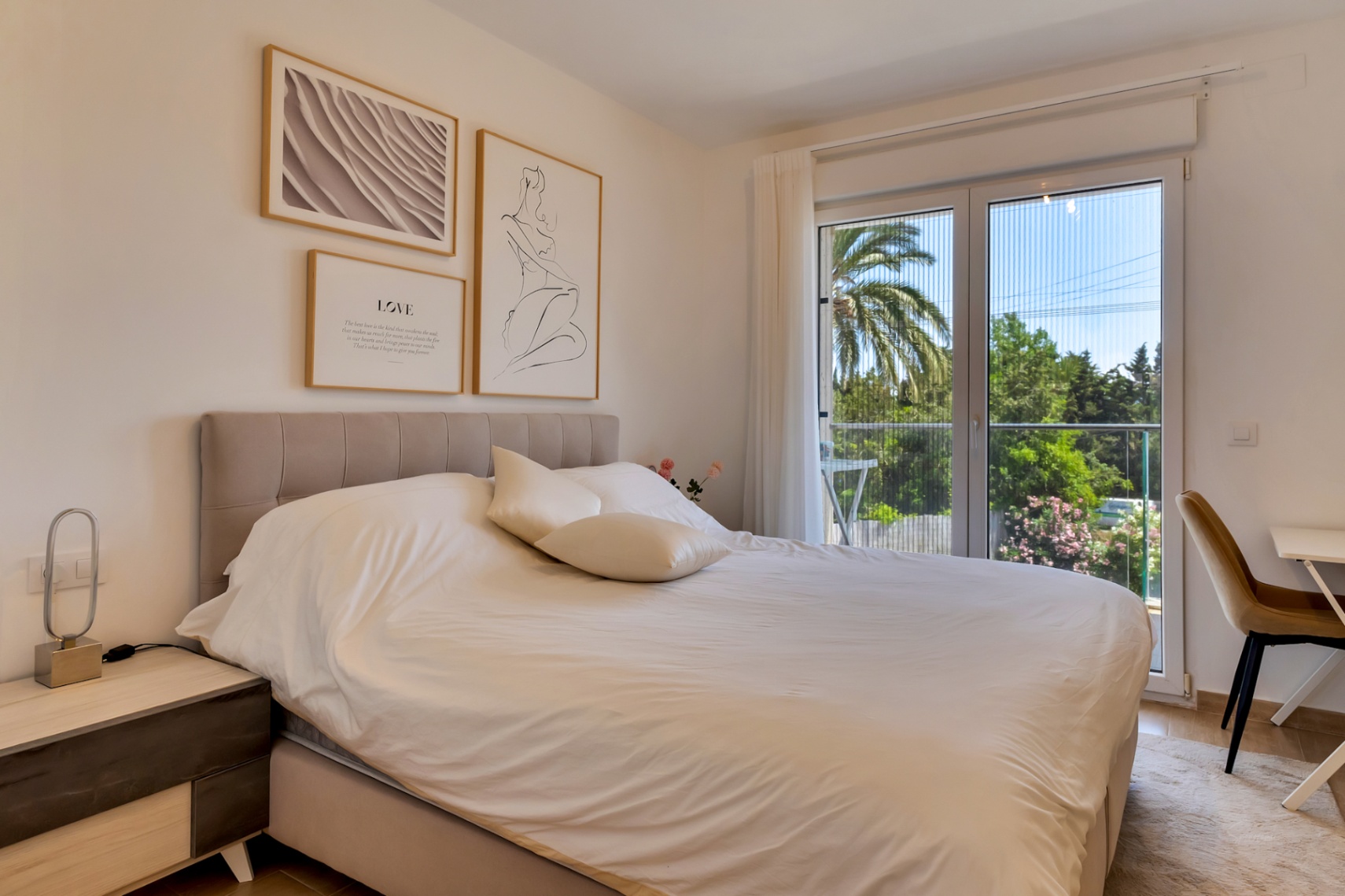 Dormitorio luminoso con cama doble, cabecero tapizado y zona de escritorio. Las puertas acristaladas con mosquitera dan acceso a un balcón privado con vistas a la vegetación.