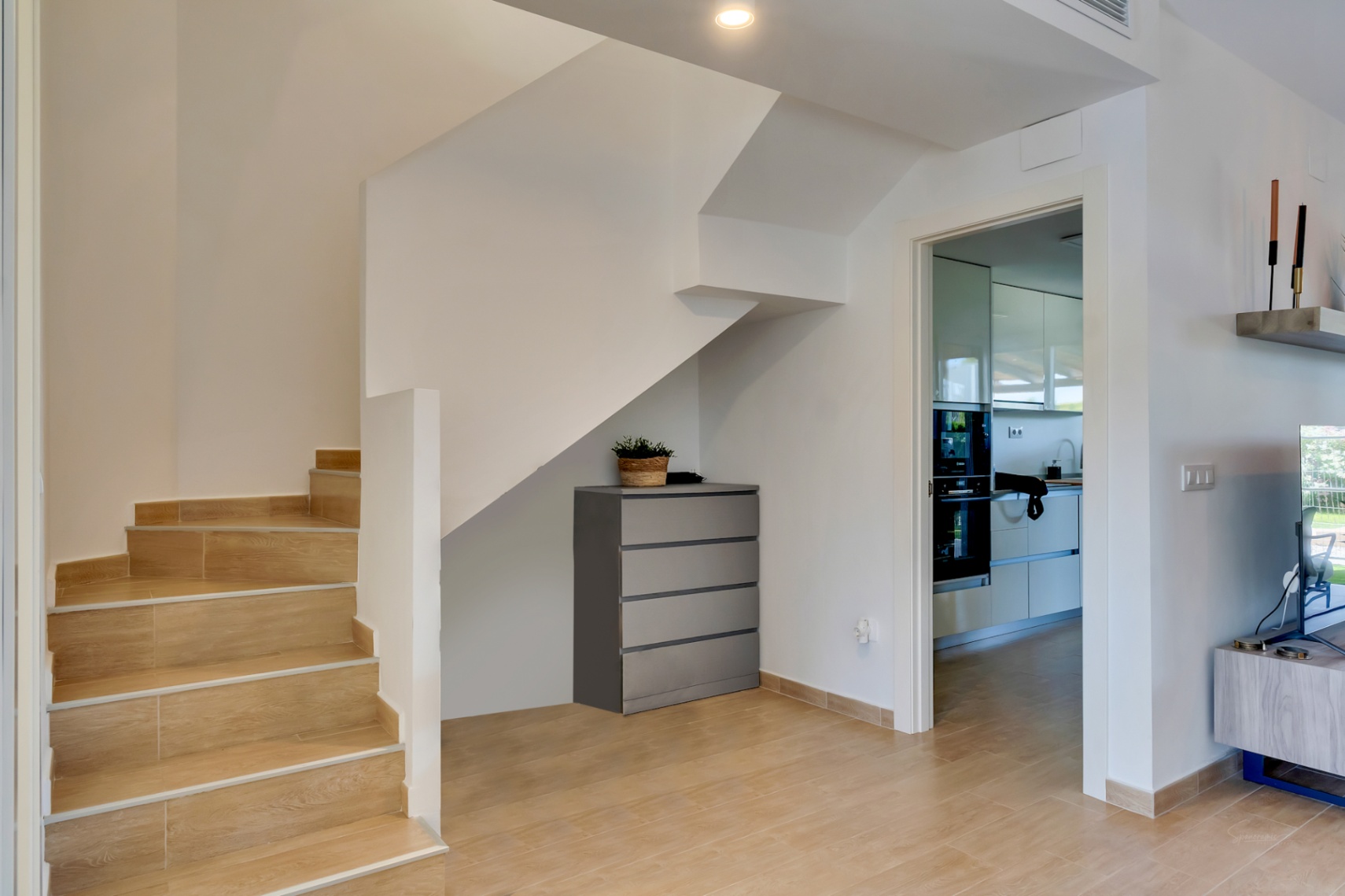 Escalera interior con peldaños de gres efecto madera y paredes blancas. Se aprecia el acceso a la cocina moderna y un práctico espacio de almacenaje bajo el hueco.