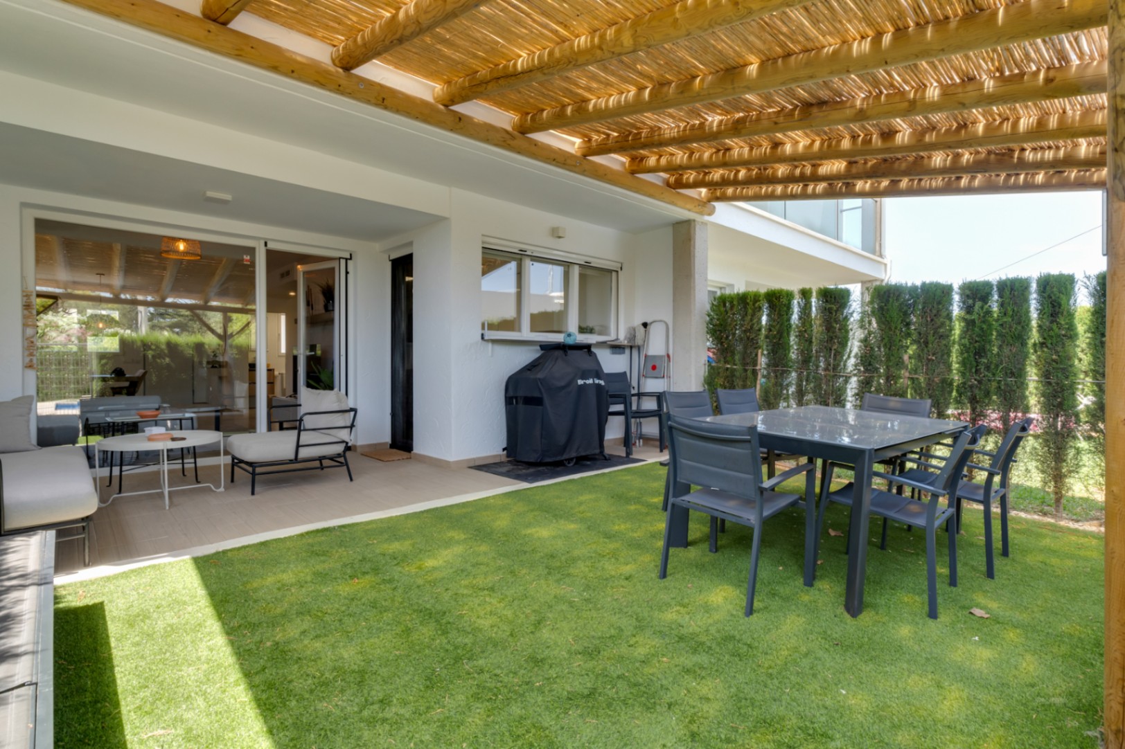 Patio privado con pérgola de madera y cañizo, zona de comedor exterior y césped artificial, conectado al interior mediante grandes puertas correderas de cristal.