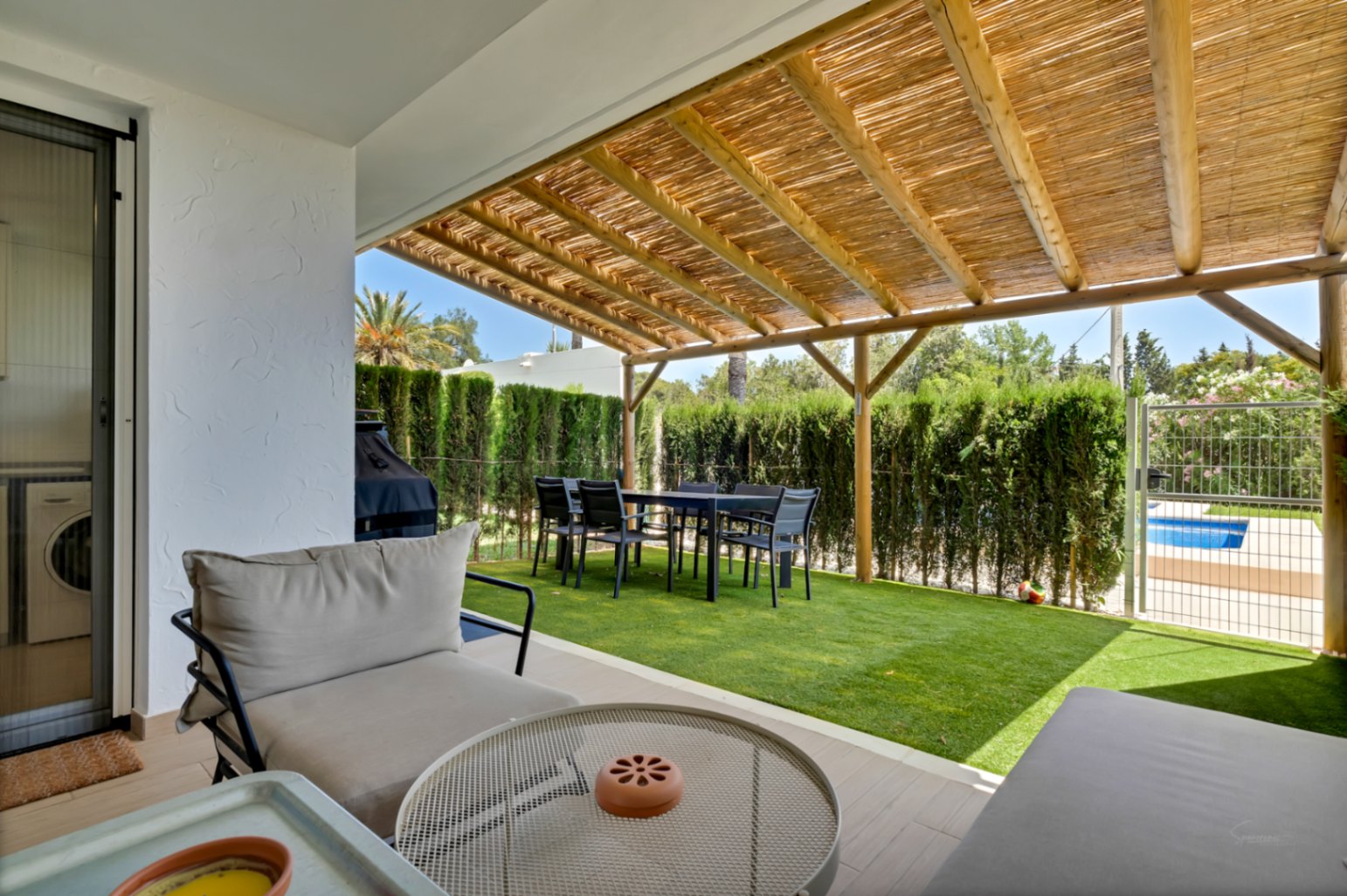 Patio privado con pérgola de madera y cañizo, zona de comedor exterior y césped artificial. Dispone de acceso directo a la piscina comunitaria mediante una puerta trasera.
