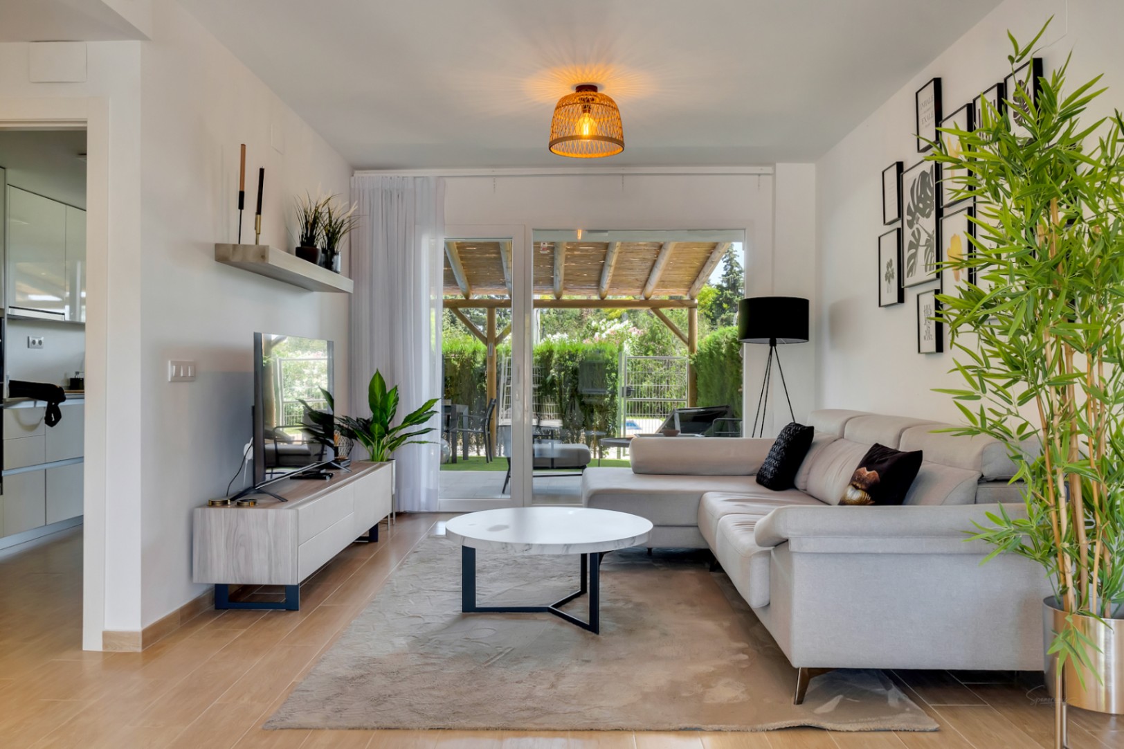 Salón luminoso con sofá en L, mesa de mármol y grandes ventanales que conectan directamente con el patio privado y su pérgola de madera, bajo una cálida lámpara de fibras naturales.