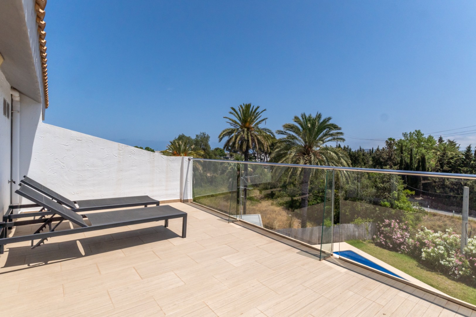 Terraza en la planta superior con suelo cerámico, barandilla de cristal y tumbonas, ofreciendo vistas despejadas a las palmeras y zonas verdes circundantes.