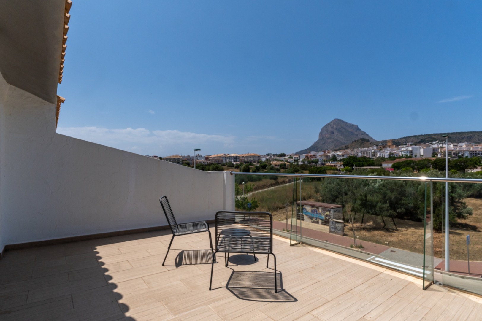 Terraza en planta superior con barandilla de cristal y vistas despejadas al Montgó y Jávea. Incluye mobiliario de exterior minimalista sobre suelo cerámico claro.