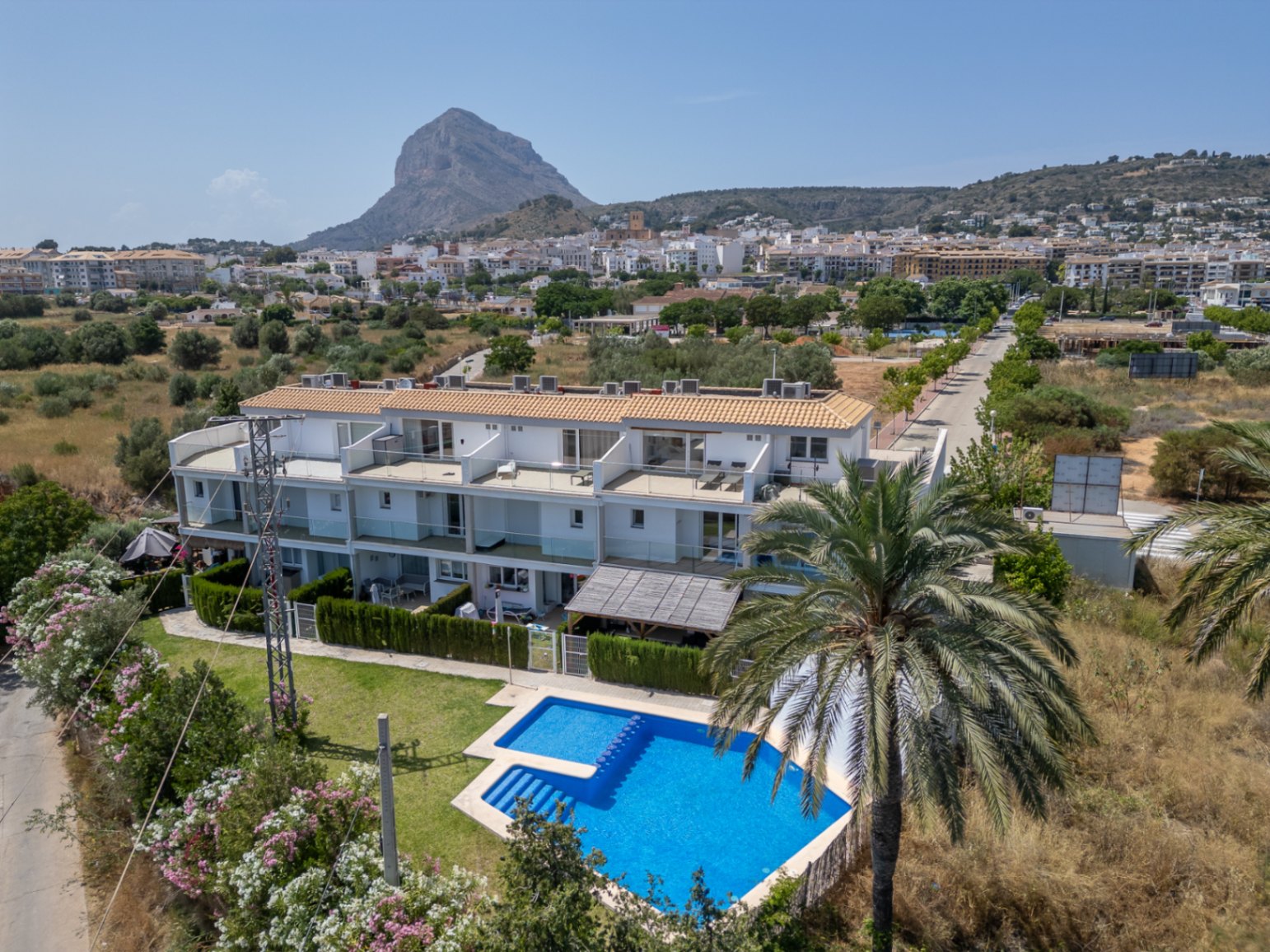 Vista aérea del complejo residencial con piscina comunitaria, jardines y vistas despejadas al Montgó y al casco antiguo de Jávea.