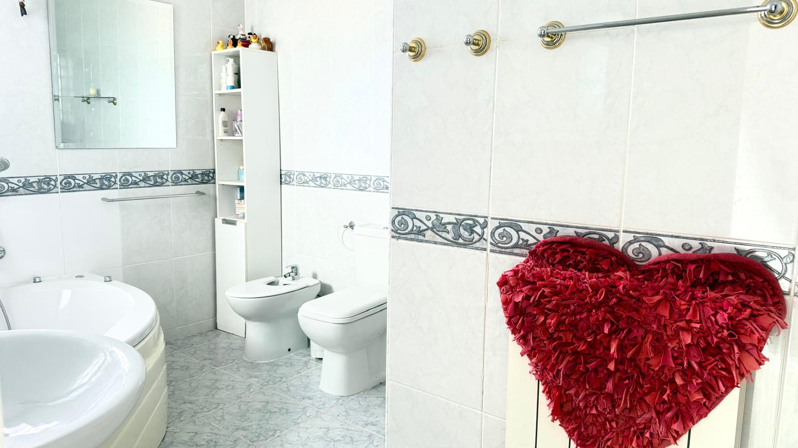 Baño completo con bañera de hidromasaje en esquina, sanitarios blancos y paredes alicatadas con cenefa decorativa. Incluye bidet y mobiliario de almacenaje vertical.