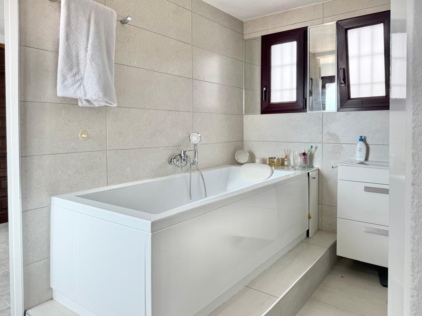 Baño con bañera blanca y reposacabezas, paredes revestidas en gres cerámico de tonos neutros y mueble de lavabo blanco junto a ventanas de carpintería oscura.